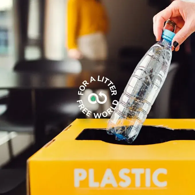 Person placing a plastic bottle into a yellow bin labeled PLASTIC for bottle recycling Person placing a plastic bottle into a yellow bin labeled PLASTIC for bottle recycling